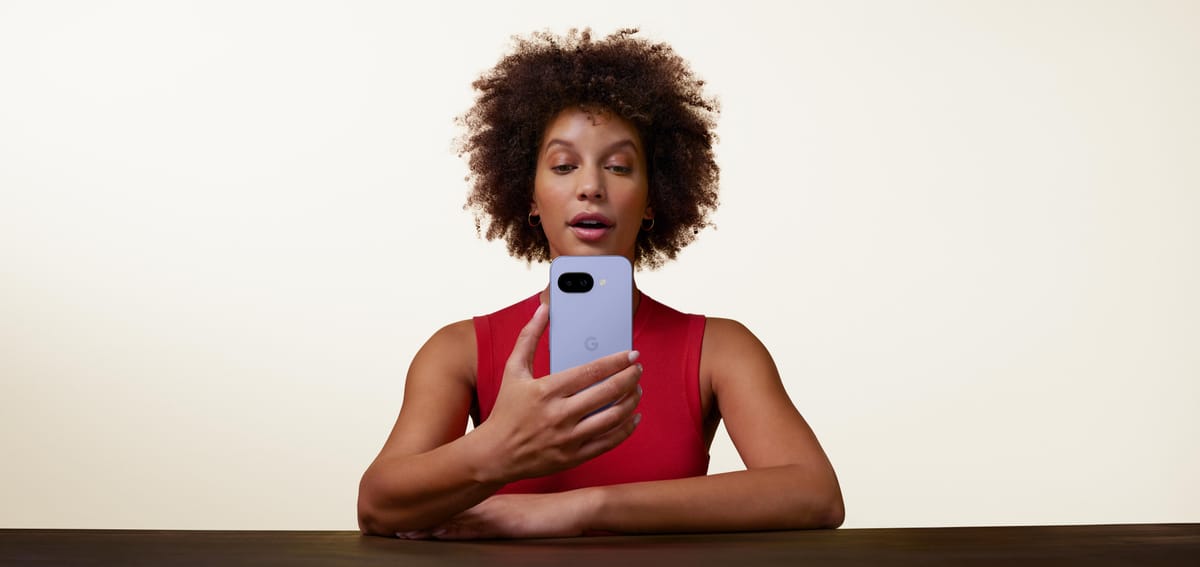 A women stares intently at her Iris coloured Google Pixel 9a smartphone. She is set against a pure white backdrop, with her arms folded leaning against a wooden table.