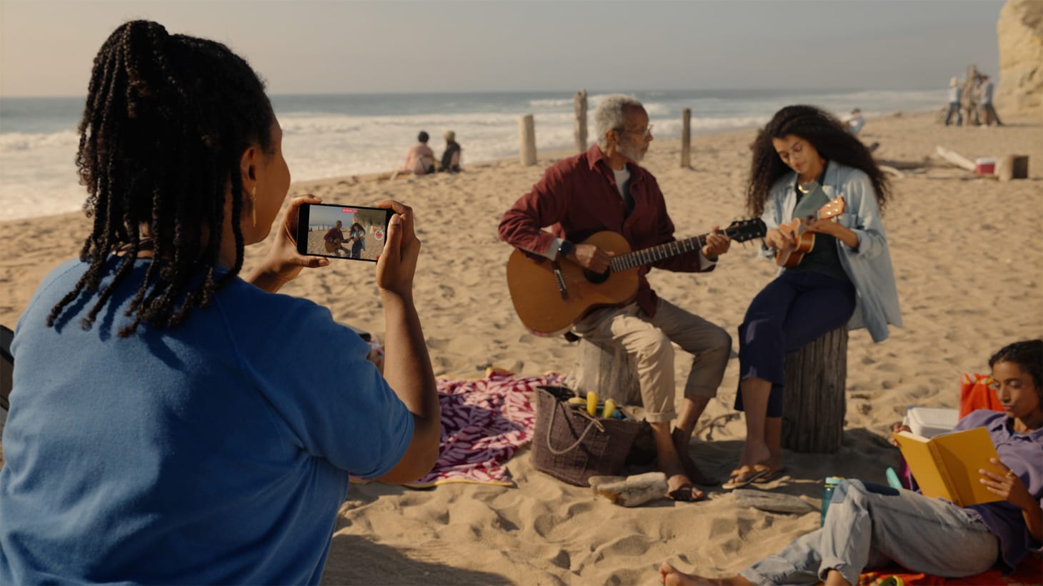 Beach scene with two guitar players while a woman videos on her iPhone.