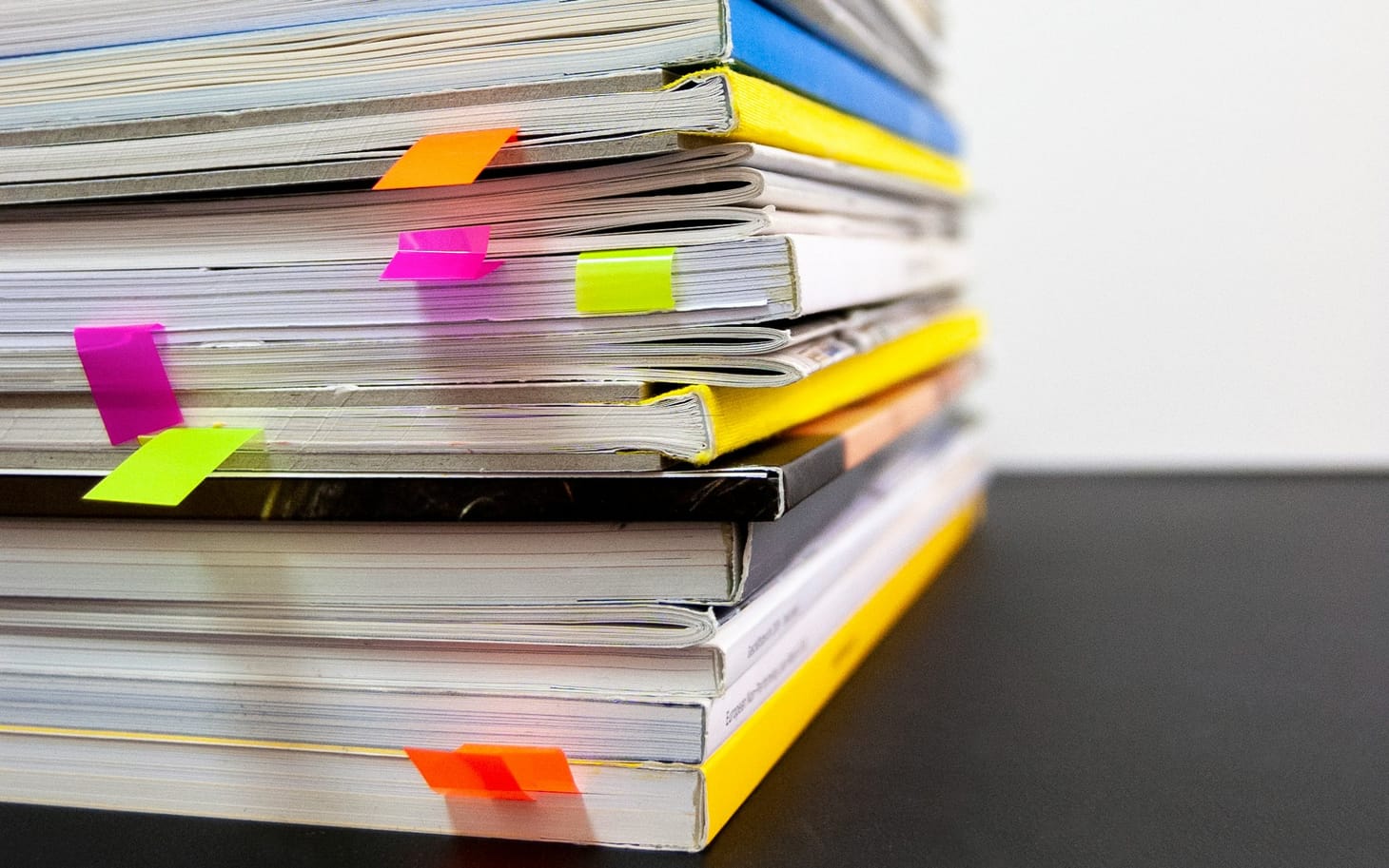 A stack of reports left of centre on a black table. Viewed low to table with stack rising out of top of frame. There are sticky tabs in various colours poking out of pages.