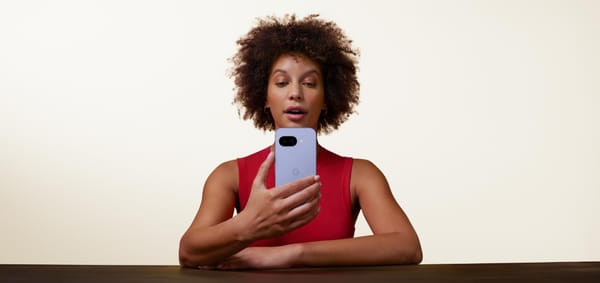 A women stares intently at her Iris coloured Google Pixel 9a smartphone. She is set against a pure white backdrop, with her arms folded leaning against a wooden table.