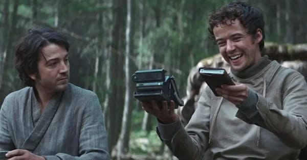 Two men sit in a forest. On the left, Andor looks with curiosity toward Nemik on the right. Nemik smiles as he holds up a notebook and an 'old' navigation device.