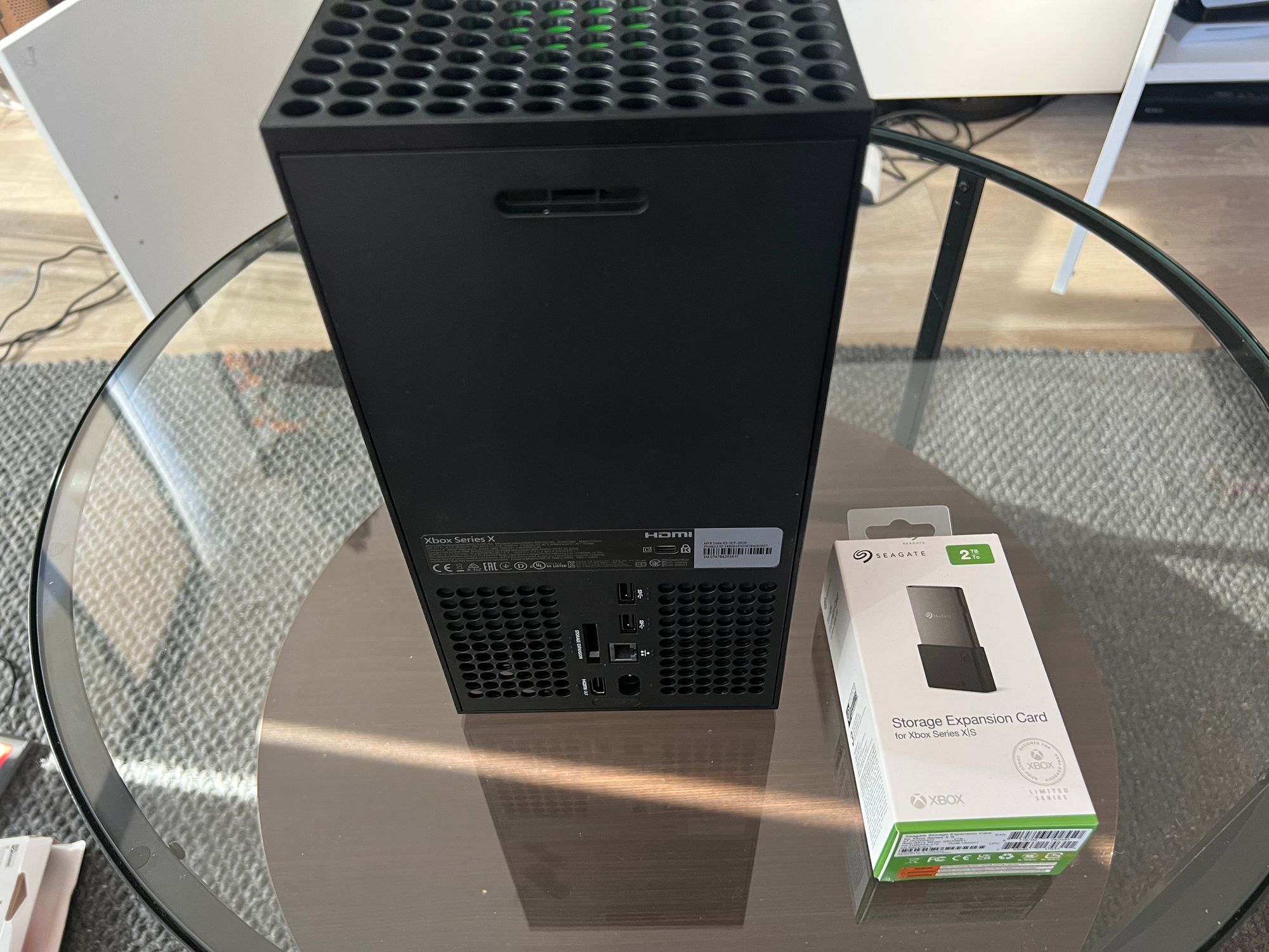 It's the same scratched glass coffee table, but this time there's a big, black, Xbox Series X on it next to a box containing a 2TB Seagate Storage Expansion Card for Xbox.